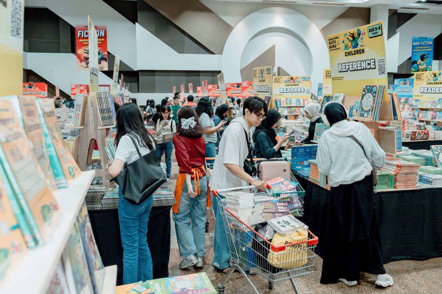 Shoppers browse colorful books and merchandise at a busy indoor book fair with shelves and a shopping cart.