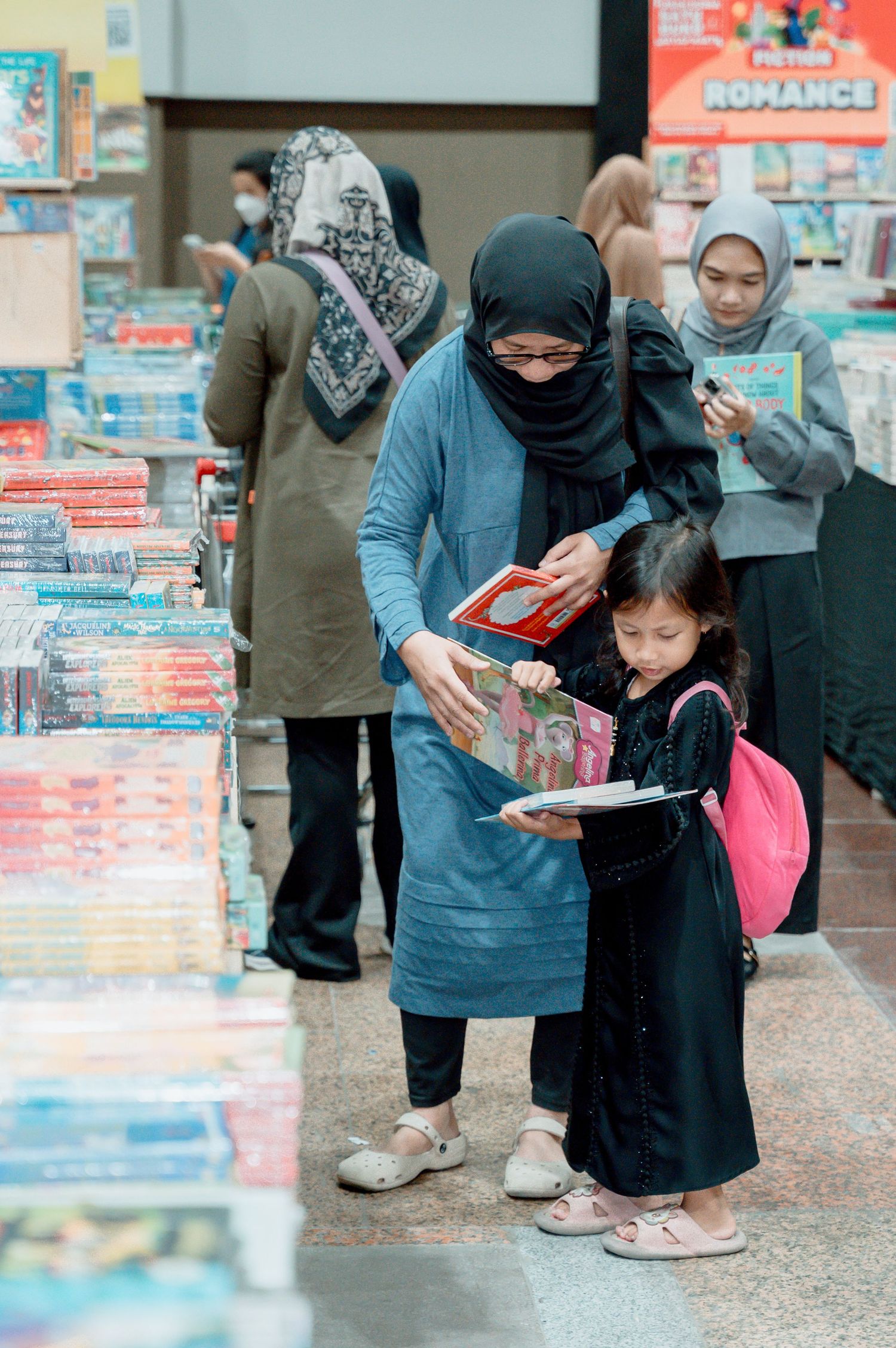 A woman in hijab helps a young girl browse books at an outdoor book fair market.