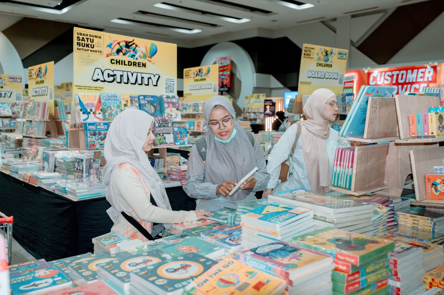 Three women in hijabs browse books at a colorful bookstore display with activity and children's books.