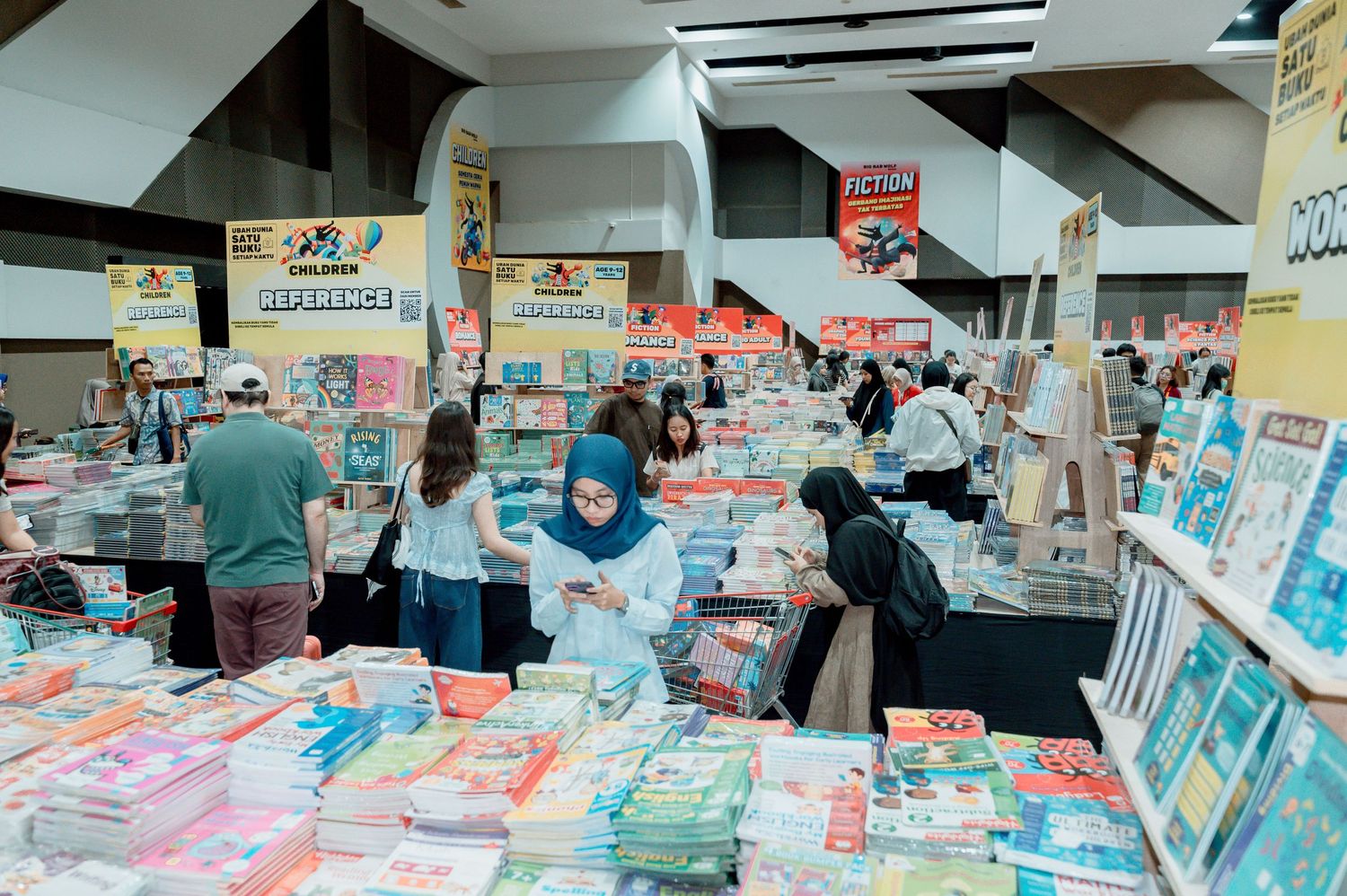 Busy book fair with shoppers browsing colorful books and educational materials displayed on tables indoors.