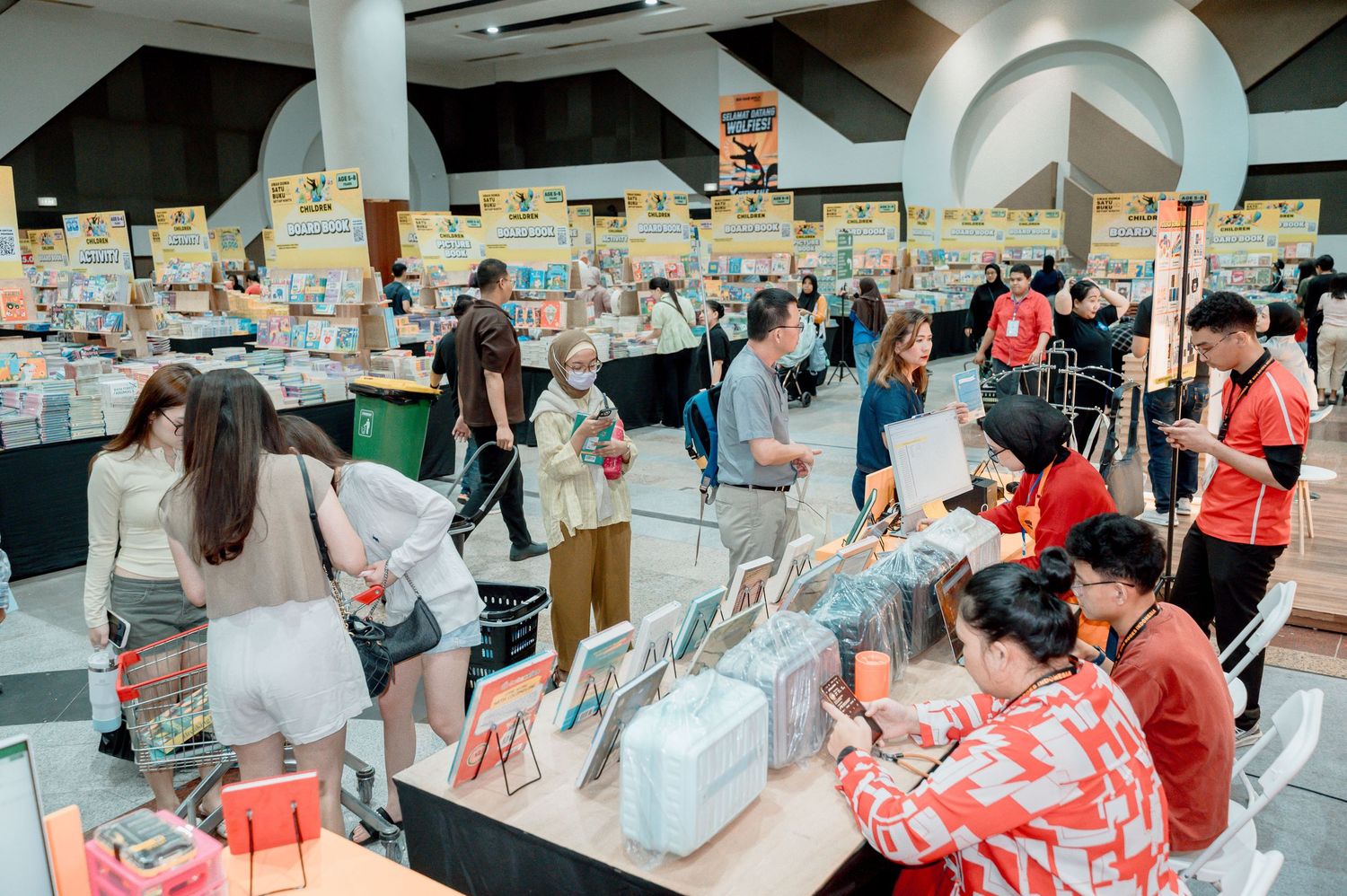 Busy book fair with shoppers browsing colorful displays of books and merchandise in a spacious indoor venue.