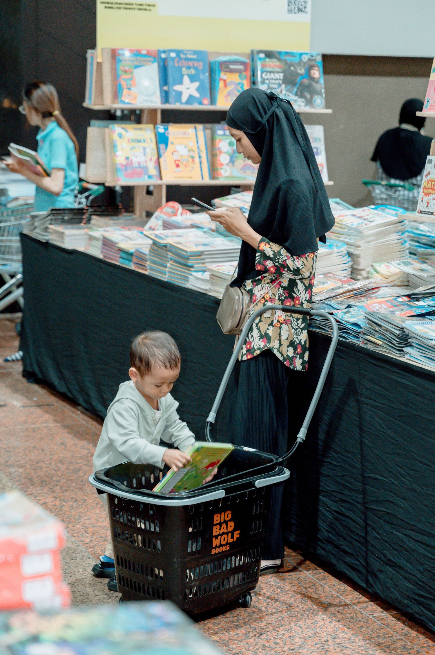 A child sits in a Big Bad Wolf shopping basket reading a book at a book fair, while a woman in hijab browses nearby.
