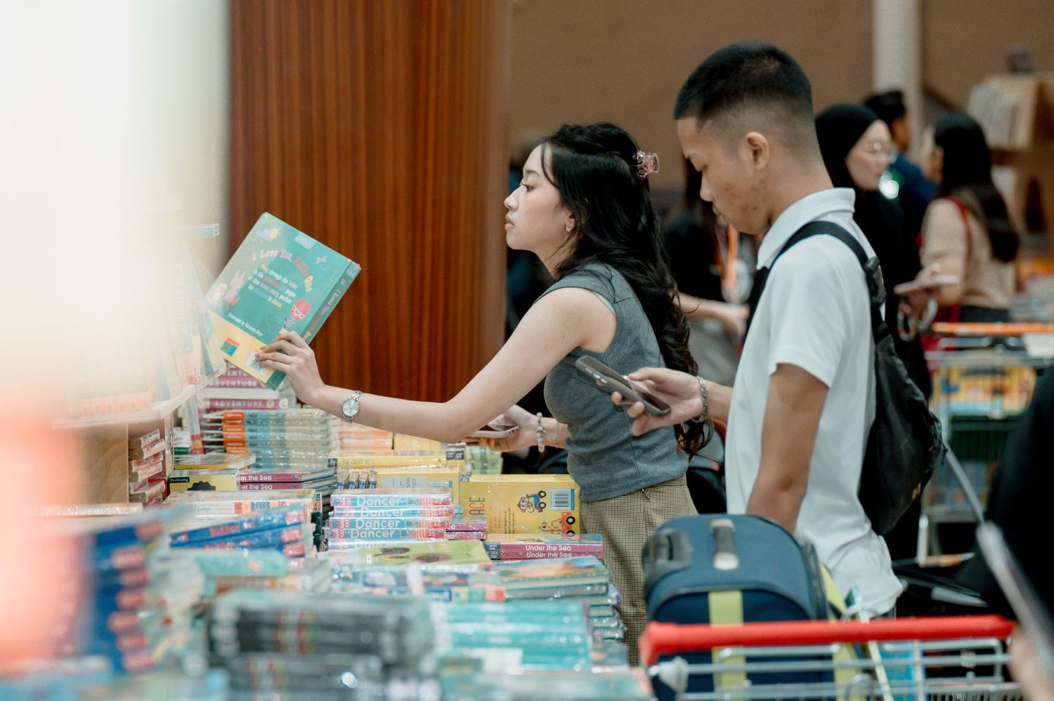 People browsing books at an indoor book fair, with a woman holding a book and a man with a backpack beside her.