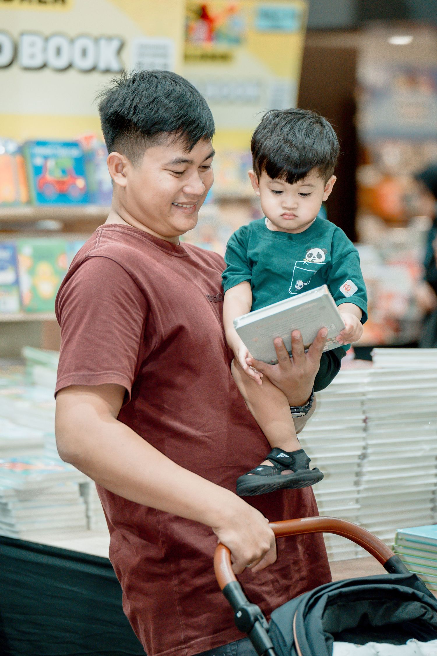 A smiling man in a red shirt holds a young boy in a green shirt while browsing books at a bookstore.