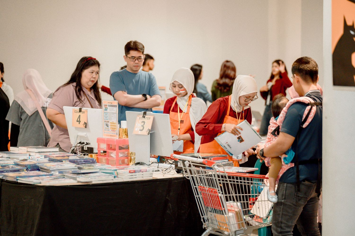 People browsing books and merchandise at an indoor market sale with a shopping cart visible.