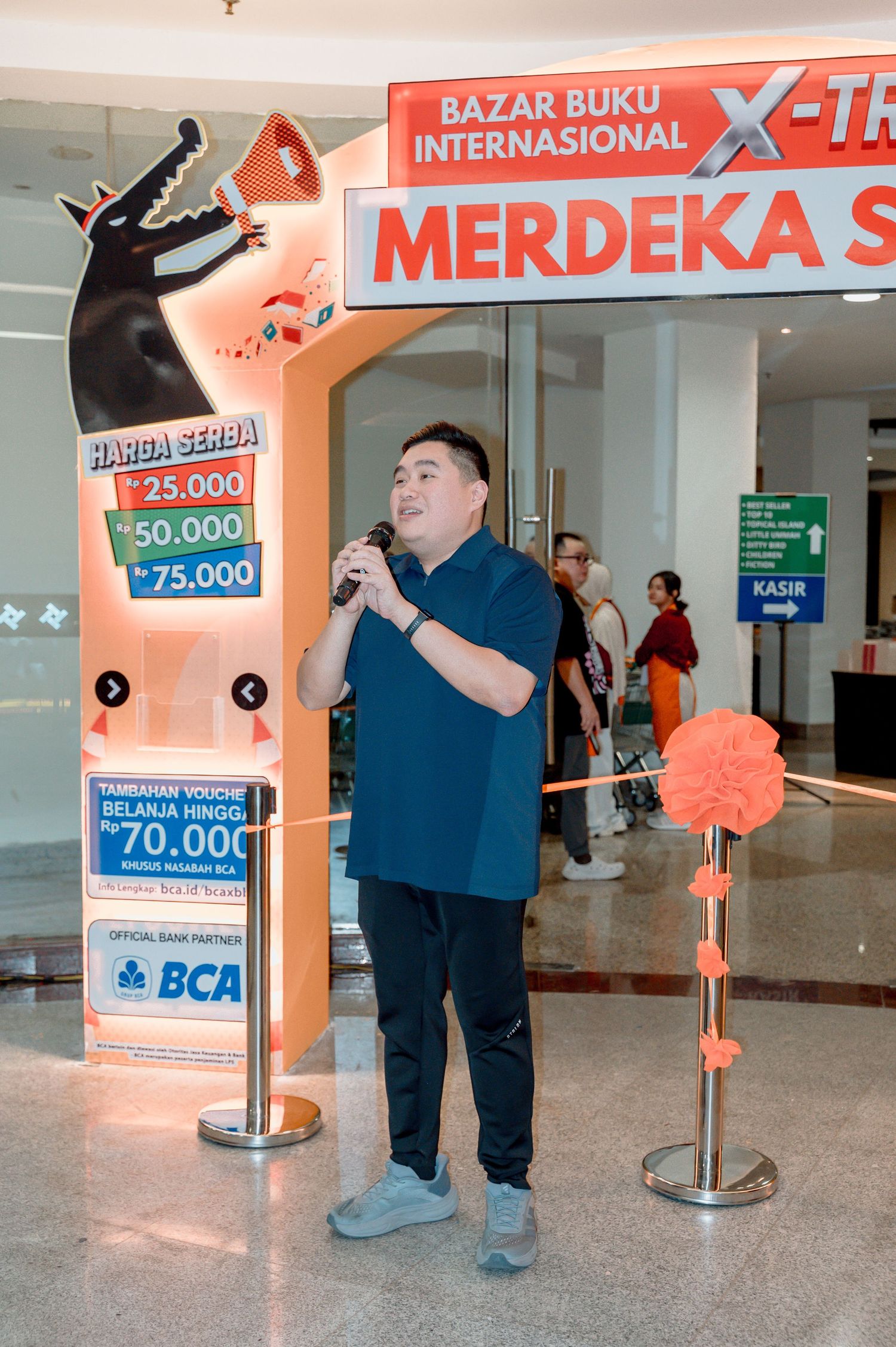 Man speaking into microphone at Bazar Buku Internasional Merdeka book fair event with BCA bank sponsorship signage.