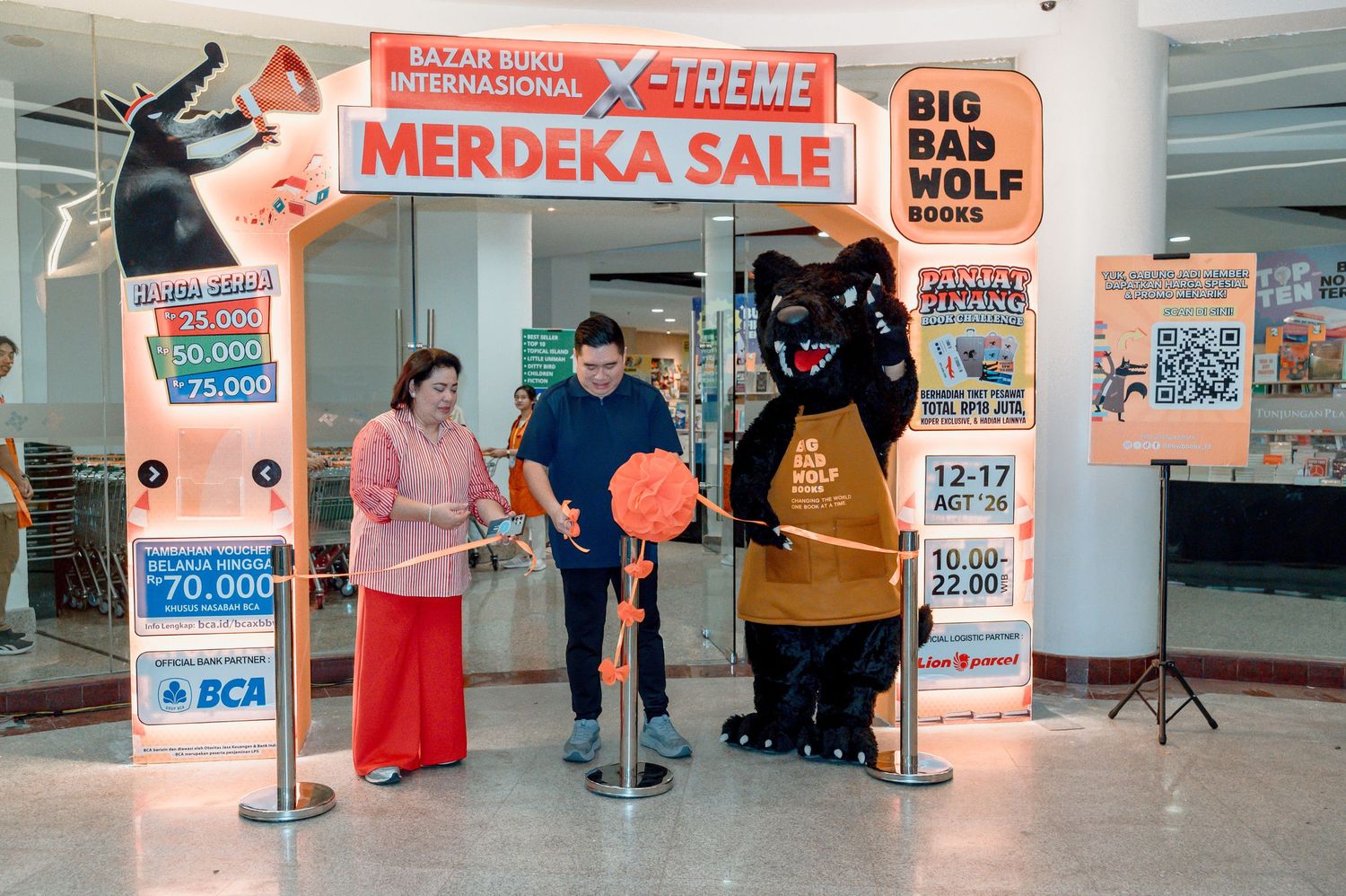 Two people and a wolf mascot cut a ribbon at the Big Bad Wolf Books X-Treme Merdeka Sale event in a mall.