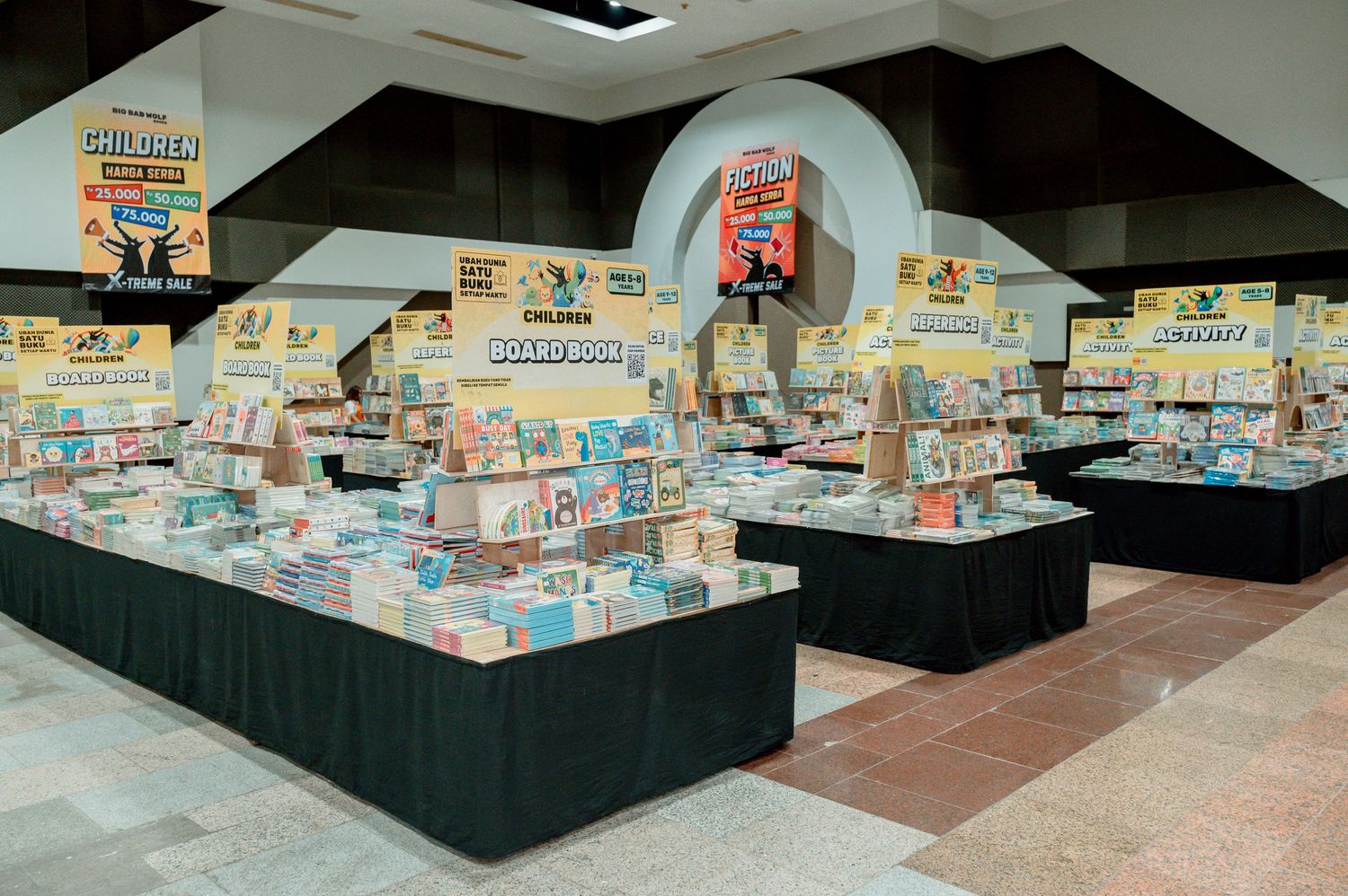 Indoor book fair display with multiple tables showcasing colorful children's books and educational materials in a large hall.