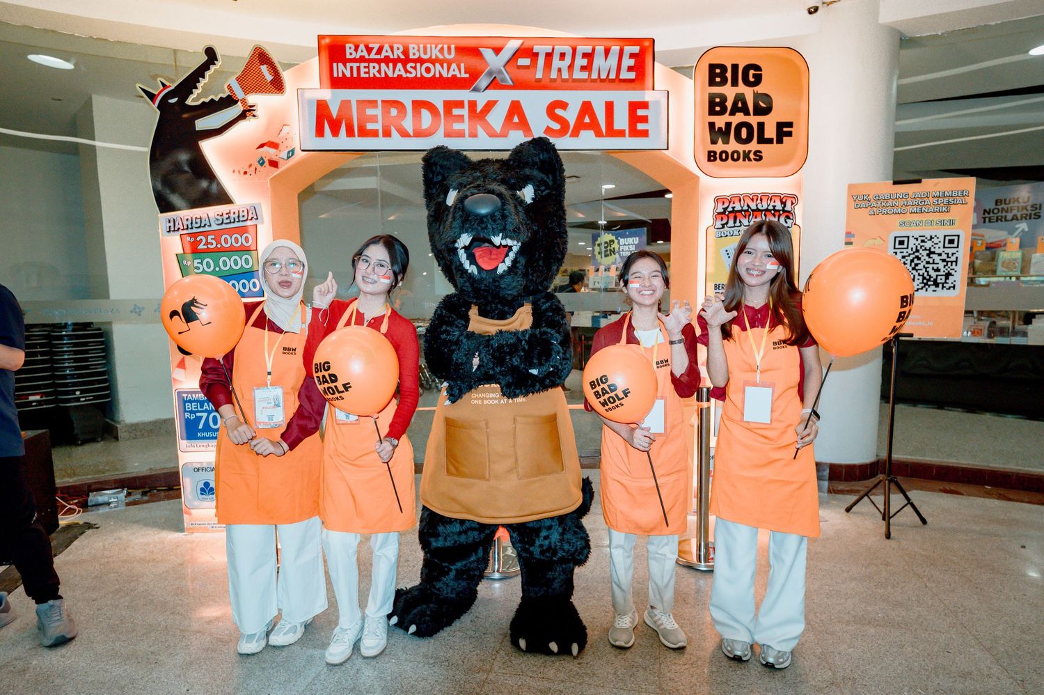 Staff and mascot pose at Big Bad Wolf Books X-Treme Merdeka Sale event with orange balloons and promotional signage.