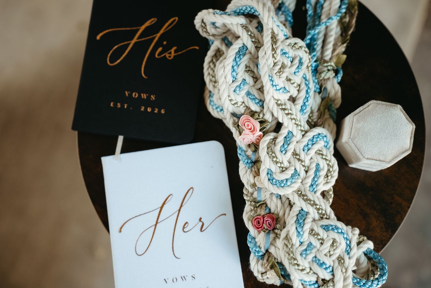 His and Her vow books beside a braided rope unity cord and ring box on a wooden table.