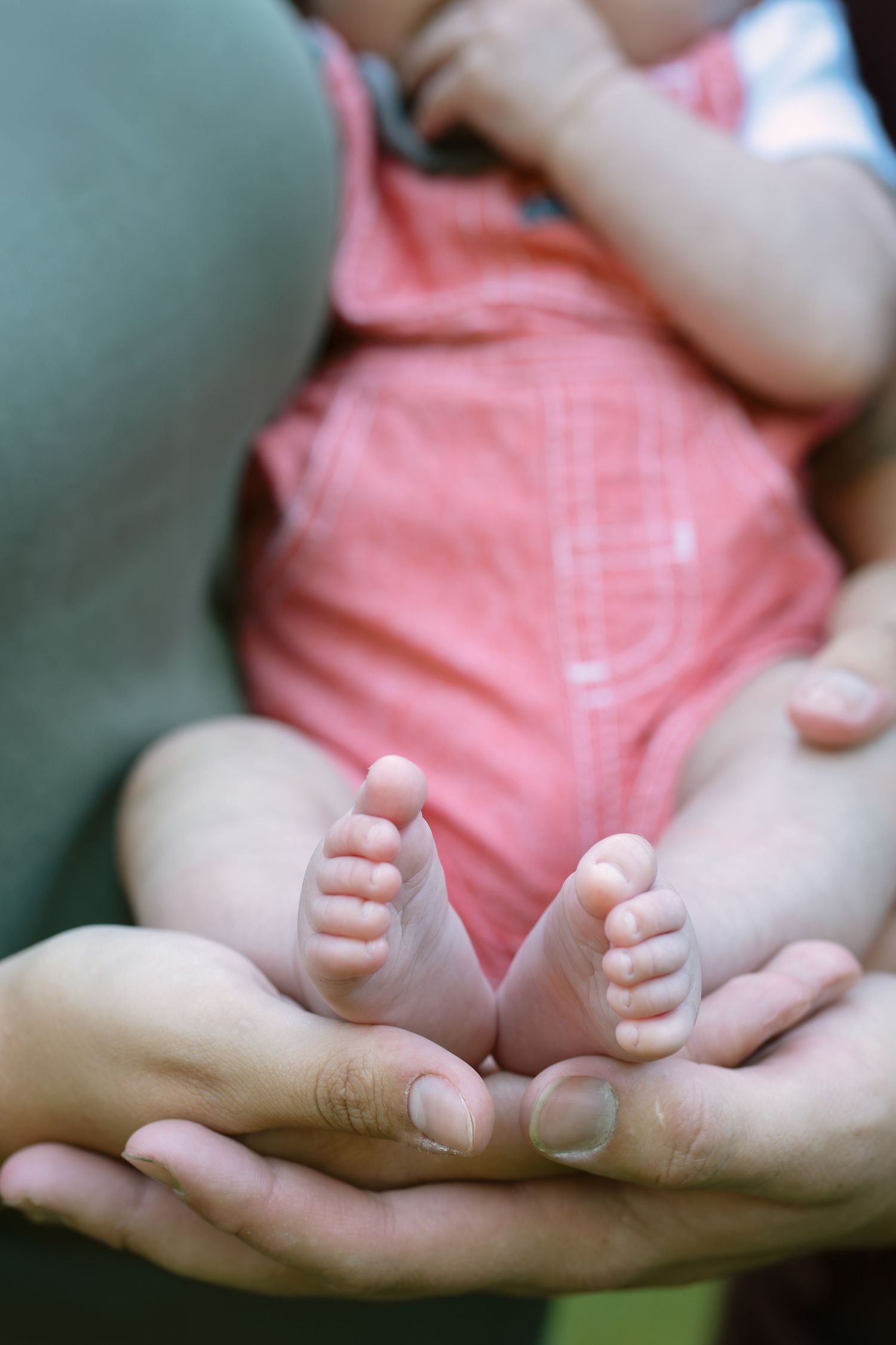 Adult hands gently cradling tiny newborn baby feet, with infant in pink onesie, close-up tender moment.