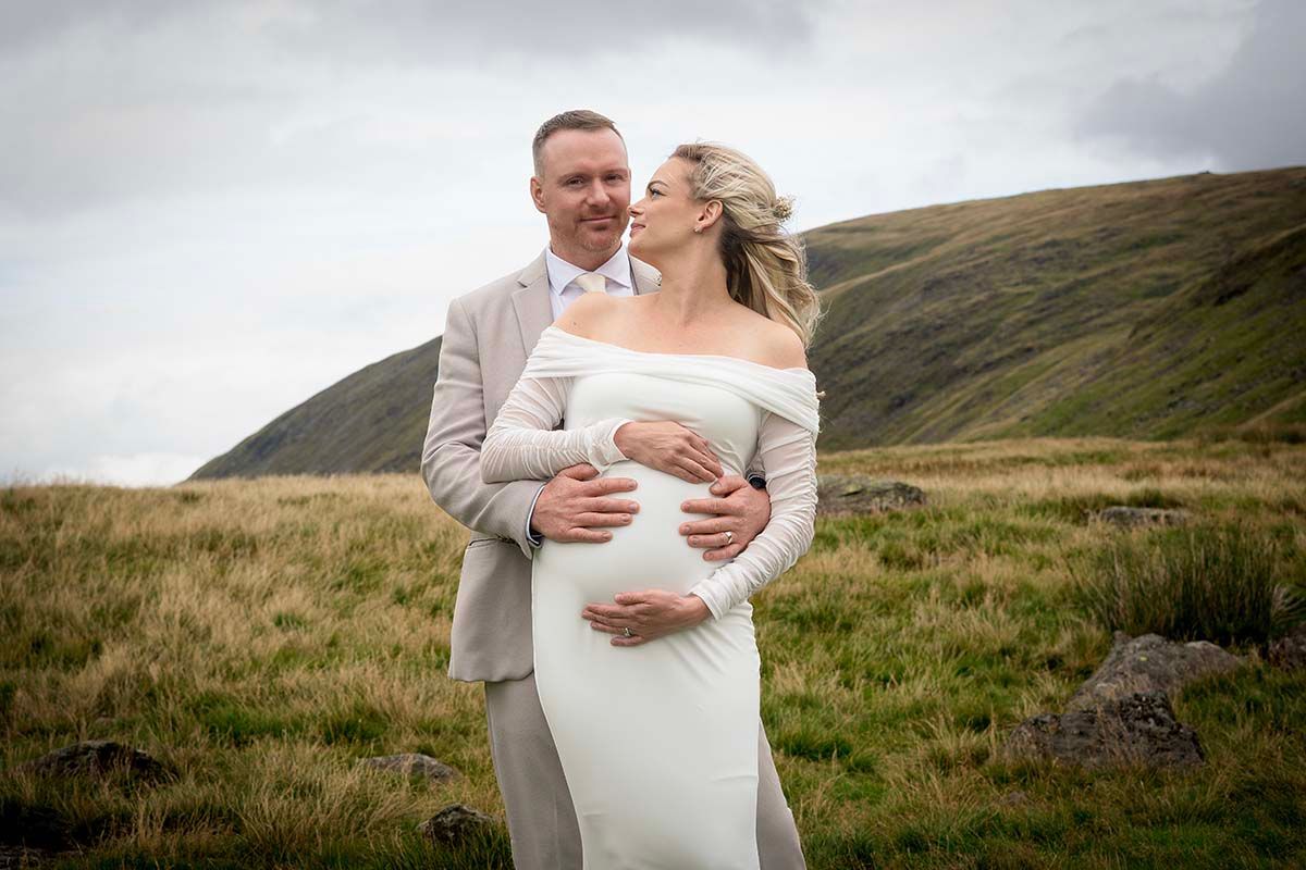 Couple in wedding attire embracing in a scenic green hillside landscape on a cloudy day during their elopement wedding at Gretna Green