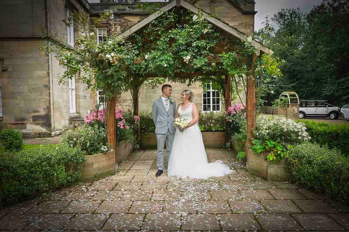 Bride and groom standing under a floral pergola at Lady Gillford's House in Carlisle, surrounded by lush gardens.