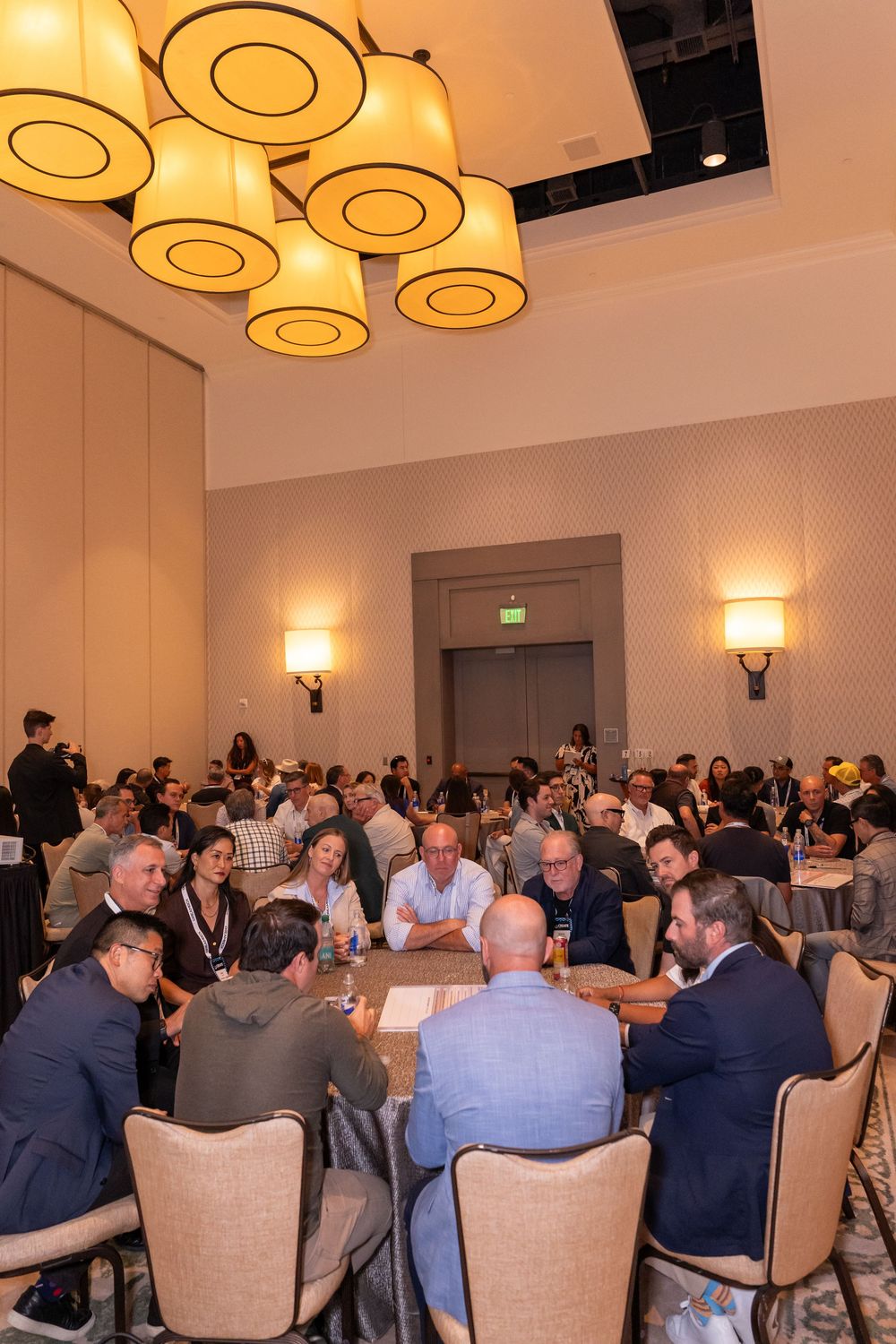 Professionals networking at round tables in an elegant conference ballroom with warm lighting and decorative pendant lamps.