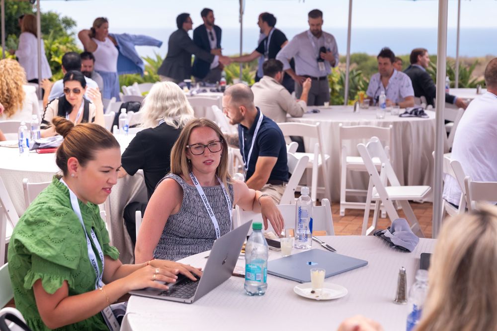 Two women collaborating on a laptop at an outdoor business conference with ocean views and white chairs.