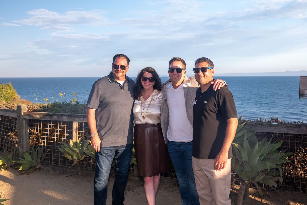 Four friends smiling together outdoors near a coastal overlook with ocean views and blue sky backdrop.