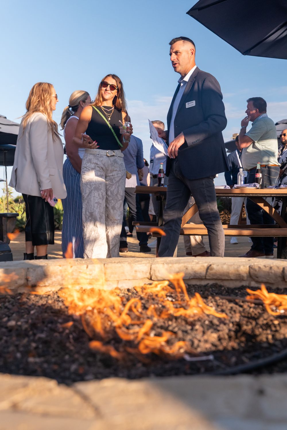 Guests mingle around an outdoor fire pit at an evening networking event under a clear blue sky.