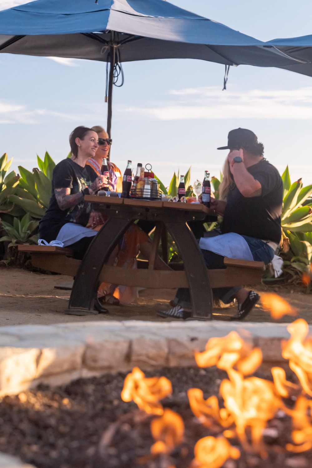 Three friends enjoy drinks at a picnic table under a patio umbrella near a glowing fire pit outdoors.