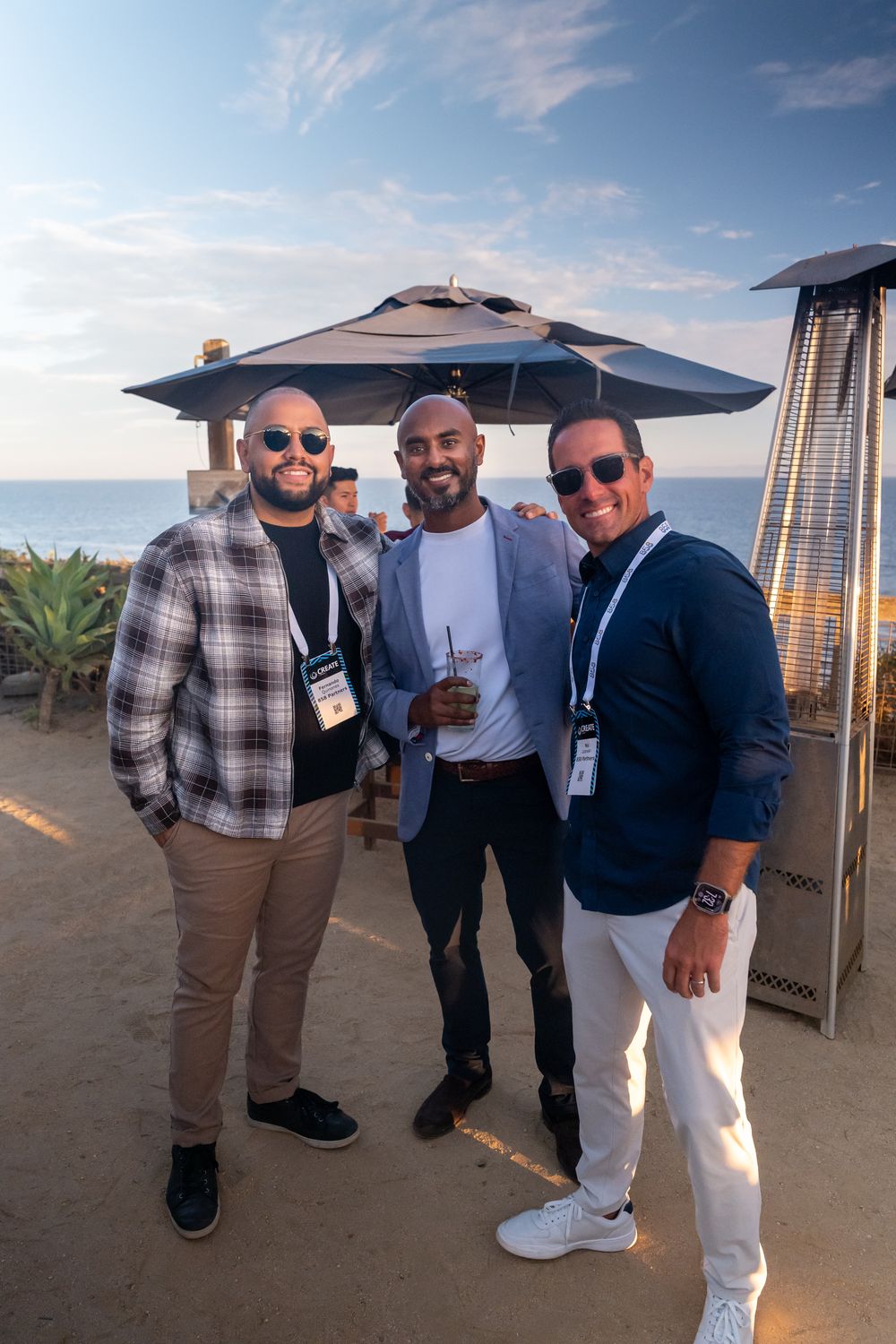 Three men smiling and posing together at an outdoor oceanview event under a patio umbrella at sunset.