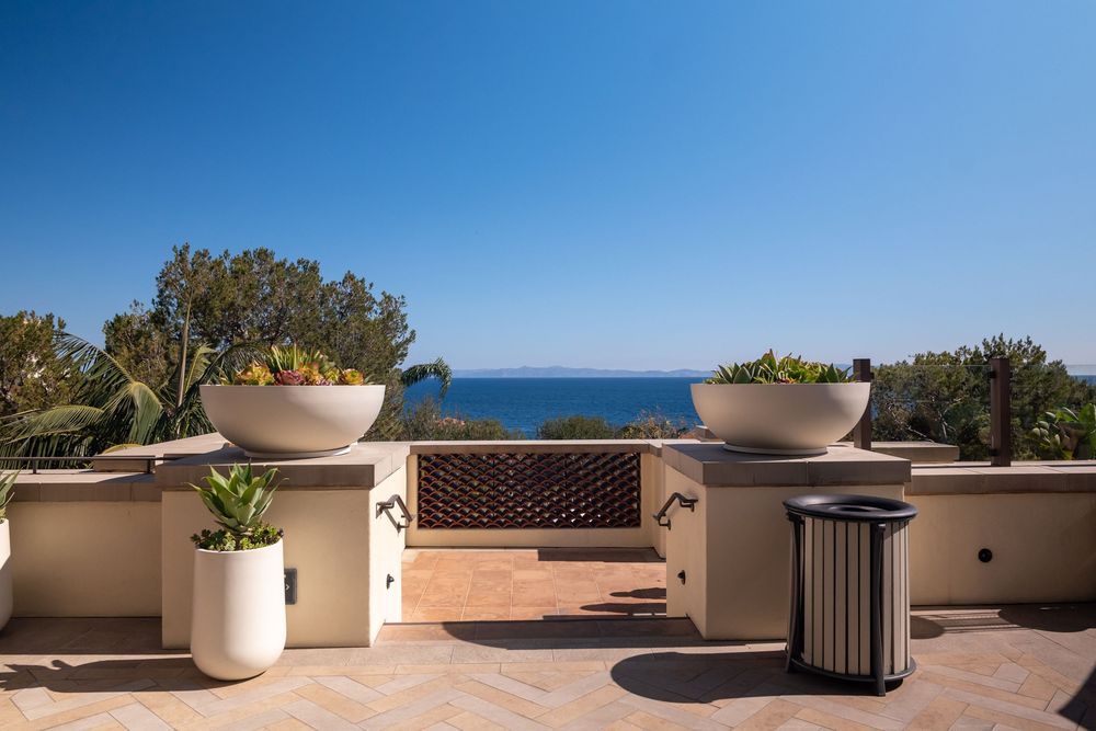 Luxury terrace with large white planters, ocean view, and decorative wine rack wall under a clear blue sky.