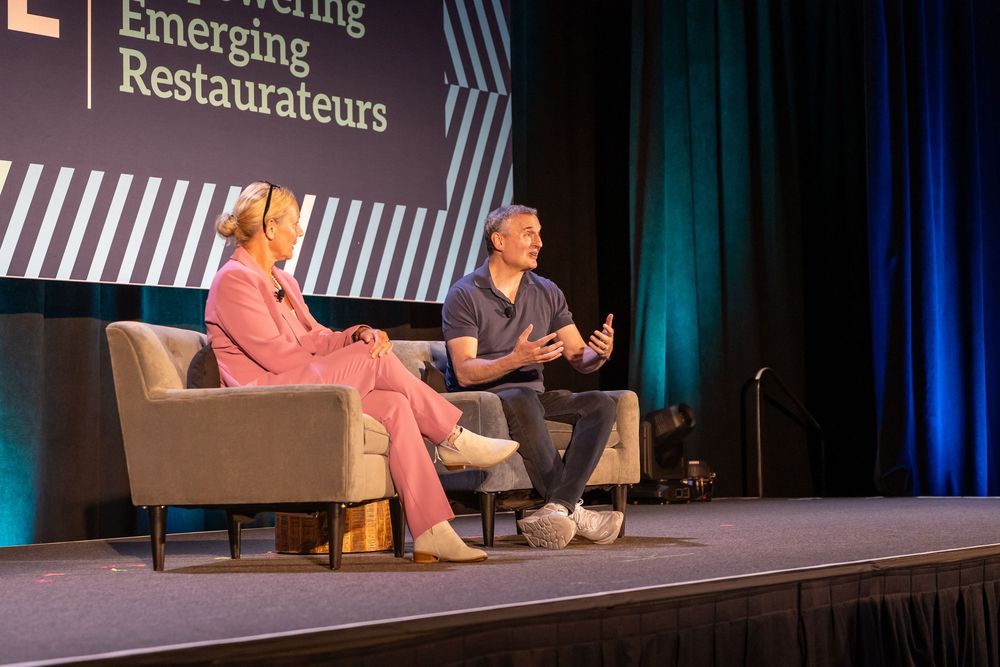 Two speakers on stage at an Empowering Emerging Restaurateurs conference, seated in chairs during a panel discussion.