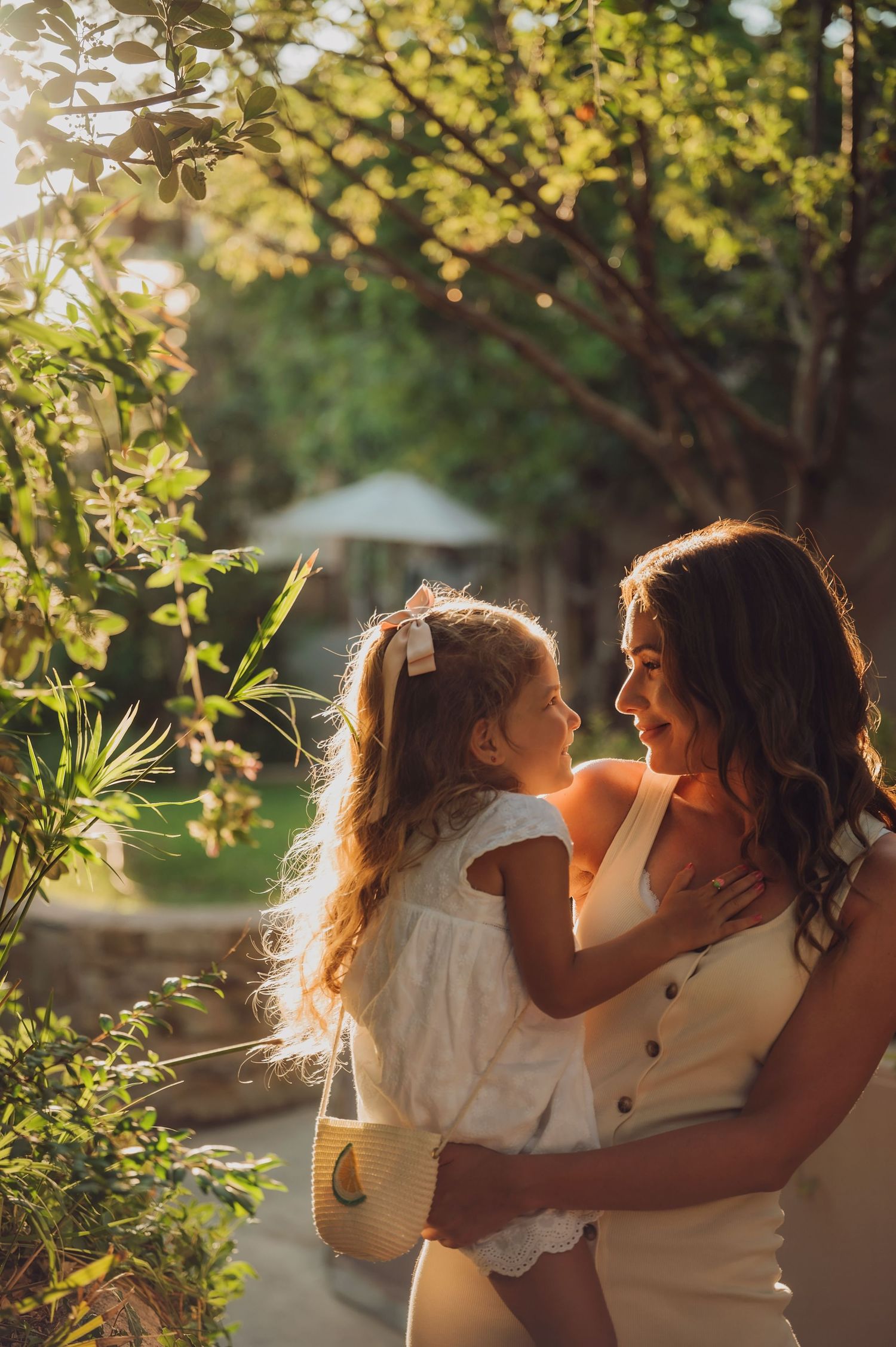 A mother holds her young daughter in a sunlit garden, both wearing white dresses and sharing a tender moment.