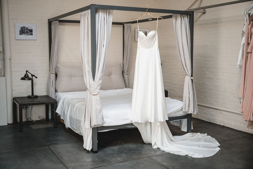 White wedding dress with a long train hanging on a four-poster bed frame in an elegant bridal suite.