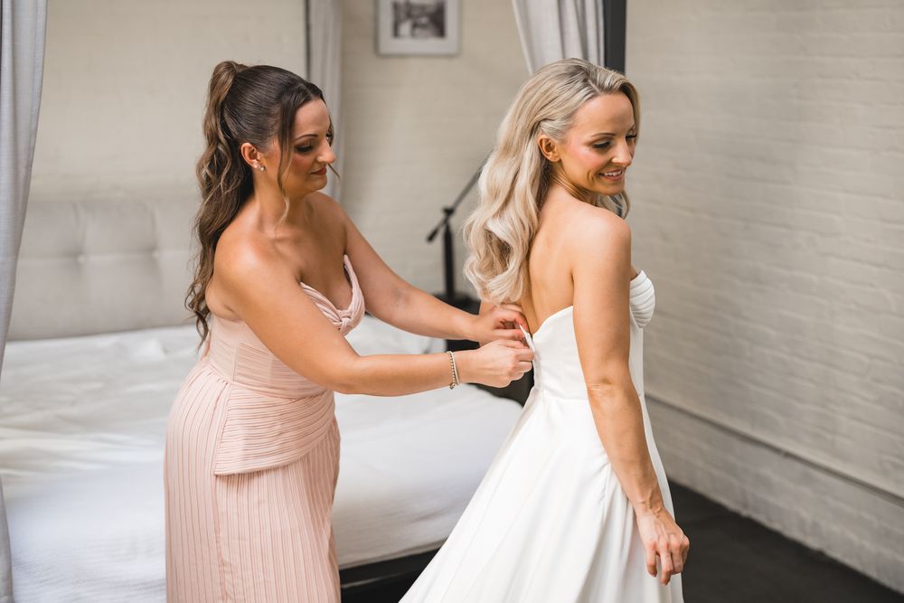 Bridesmaid in pink dress helping bride with strapless white wedding gown buttons.