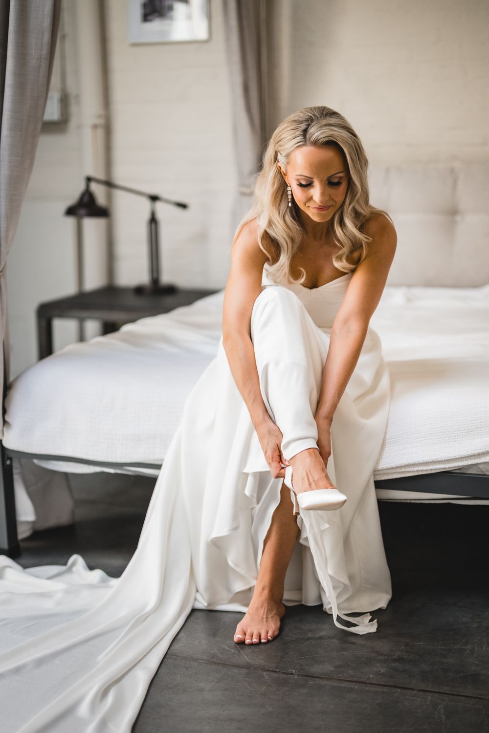 Bride in white strapless gown sitting on bed putting on heels while getting ready for her wedding day.