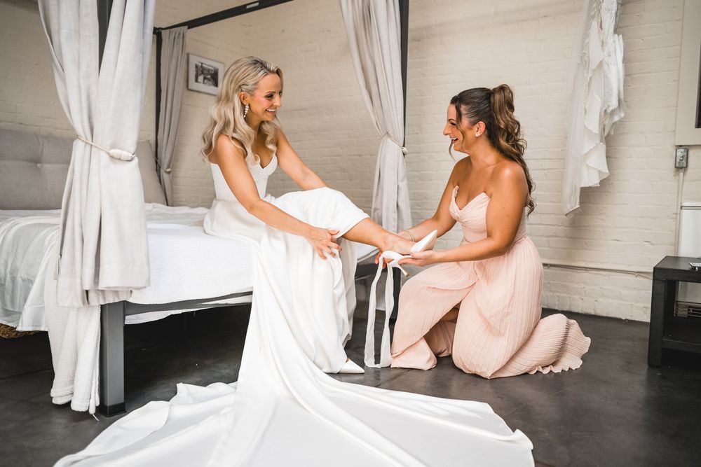 Bridesmaid kneeling on floor helping bride adjust her white wedding gown train in elegant bedroom.