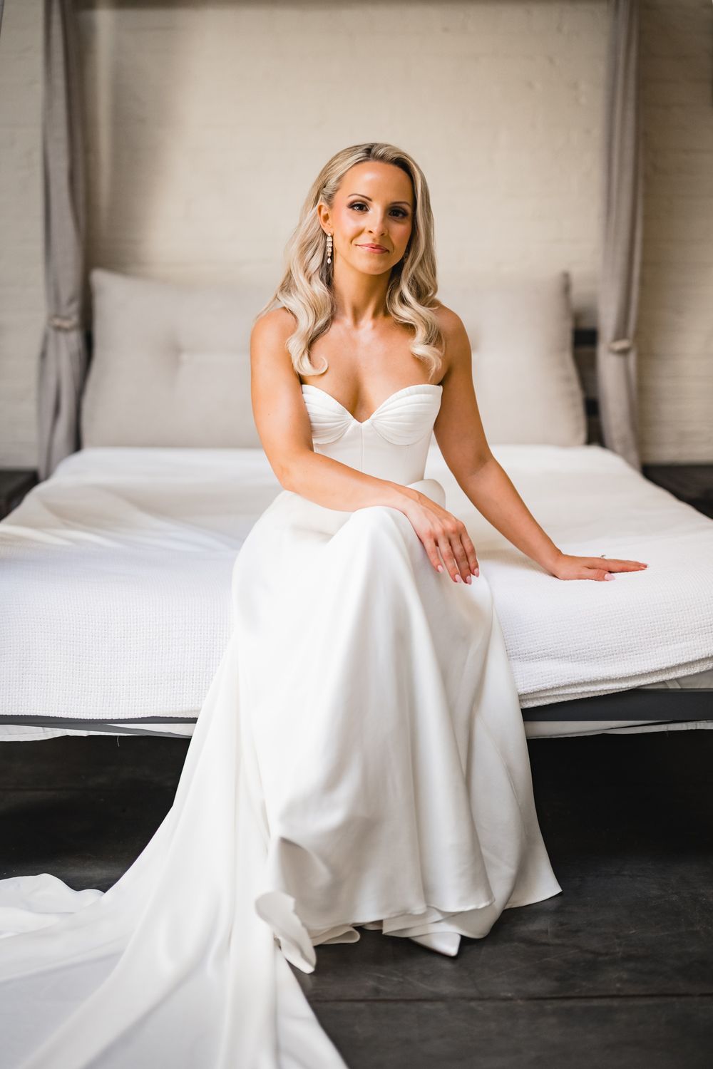 Elegant bride in strapless white ball gown sitting on bed, with flowing train and soft curled hair.