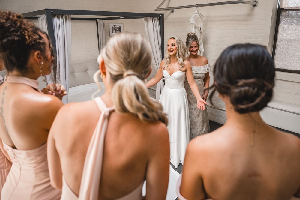 Bridesmaids in blush dresses admiring smiling bride in white wedding gown near mirror.