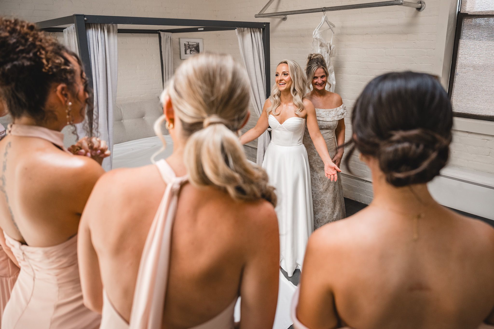 Bridesmaids in blush dresses admiring smiling bride in white wedding gown near mirror.