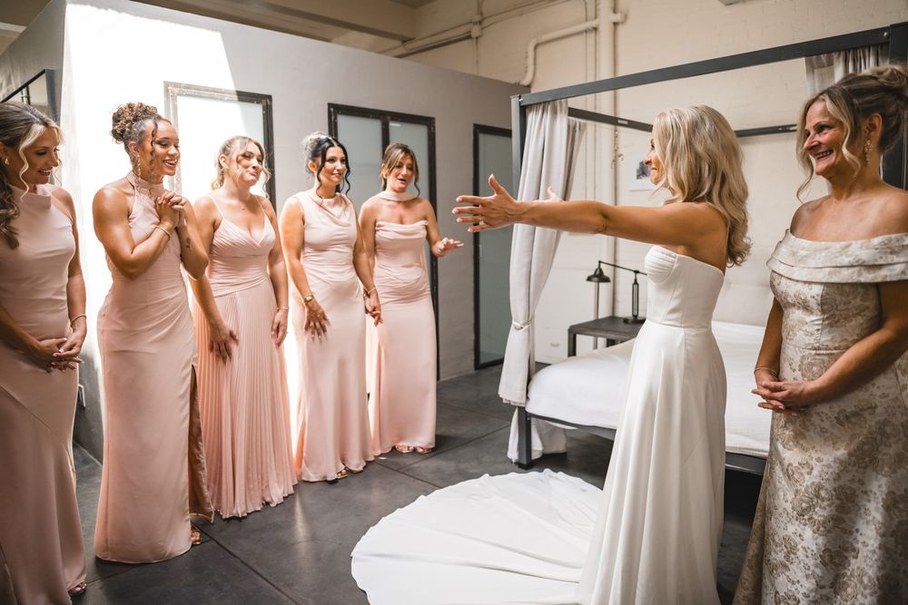 Bride in white gown revealing herself to smiling bridesmaids in blush pink dresses during wedding preparations.