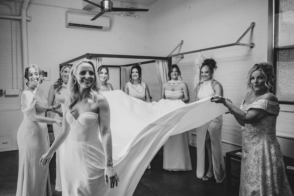 Bride and bridesmaids in white gowns spreading wedding dress train in black and white photo.