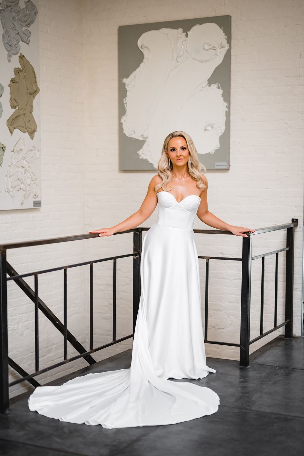 Bride in strapless white sweetheart gown with train poses gracefully at iron railing in elegant white brick venue.