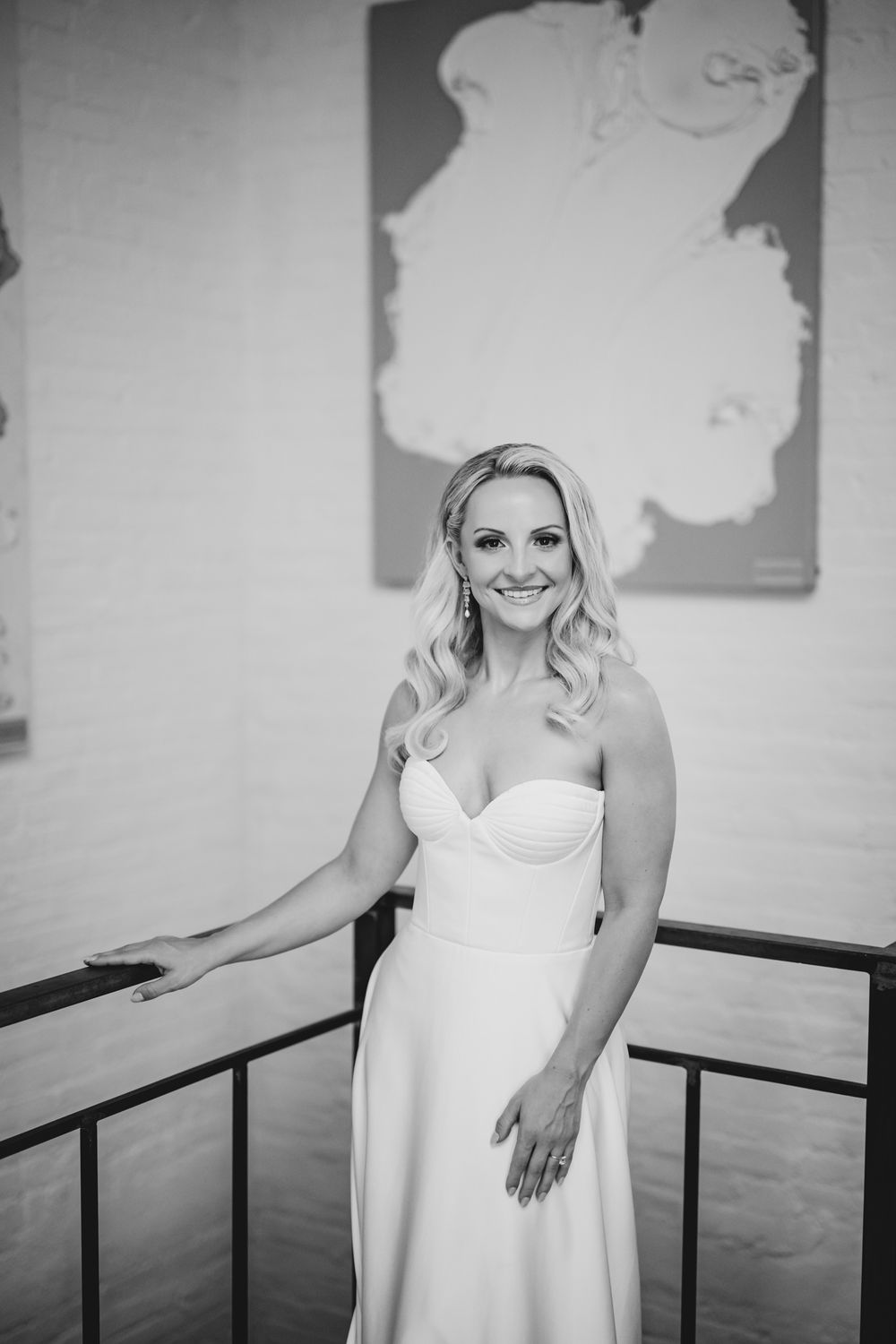 Black and white photo of a smiling bride in a strapless gown posing by a railing with a map artwork behind her.