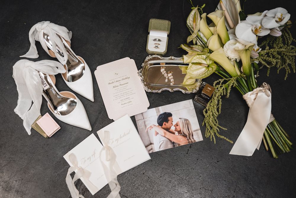 Elegant wedding flat lay featuring bridal heels, floral bouquet, invitations, a photo, ring box, and perfume on dark surface.