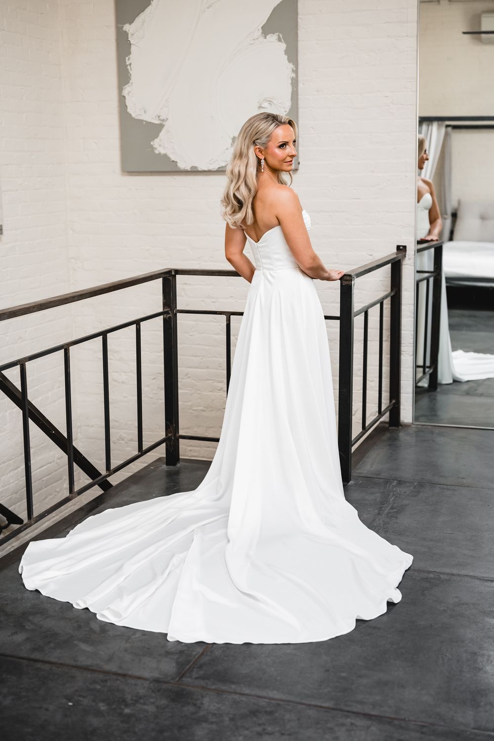 Bride in strapless white wedding gown with elegant train poses on stairs in modern venue.