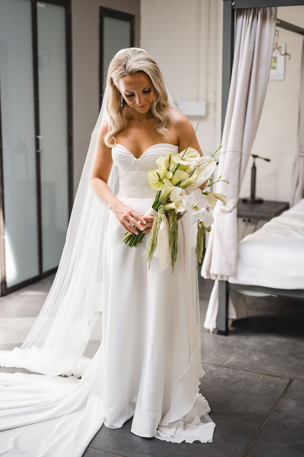 Bride in strapless white gown holding white orchid bouquet with veil, standing indoors on wedding day.