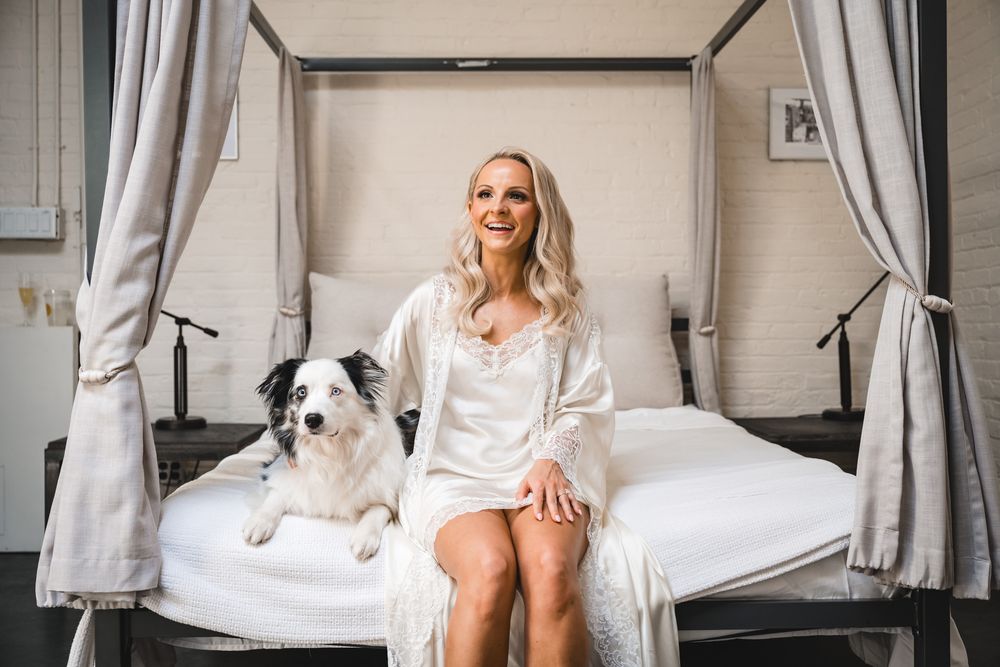 Smiling blonde woman in white robe sitting on elegant bed with black and white border collie dog.