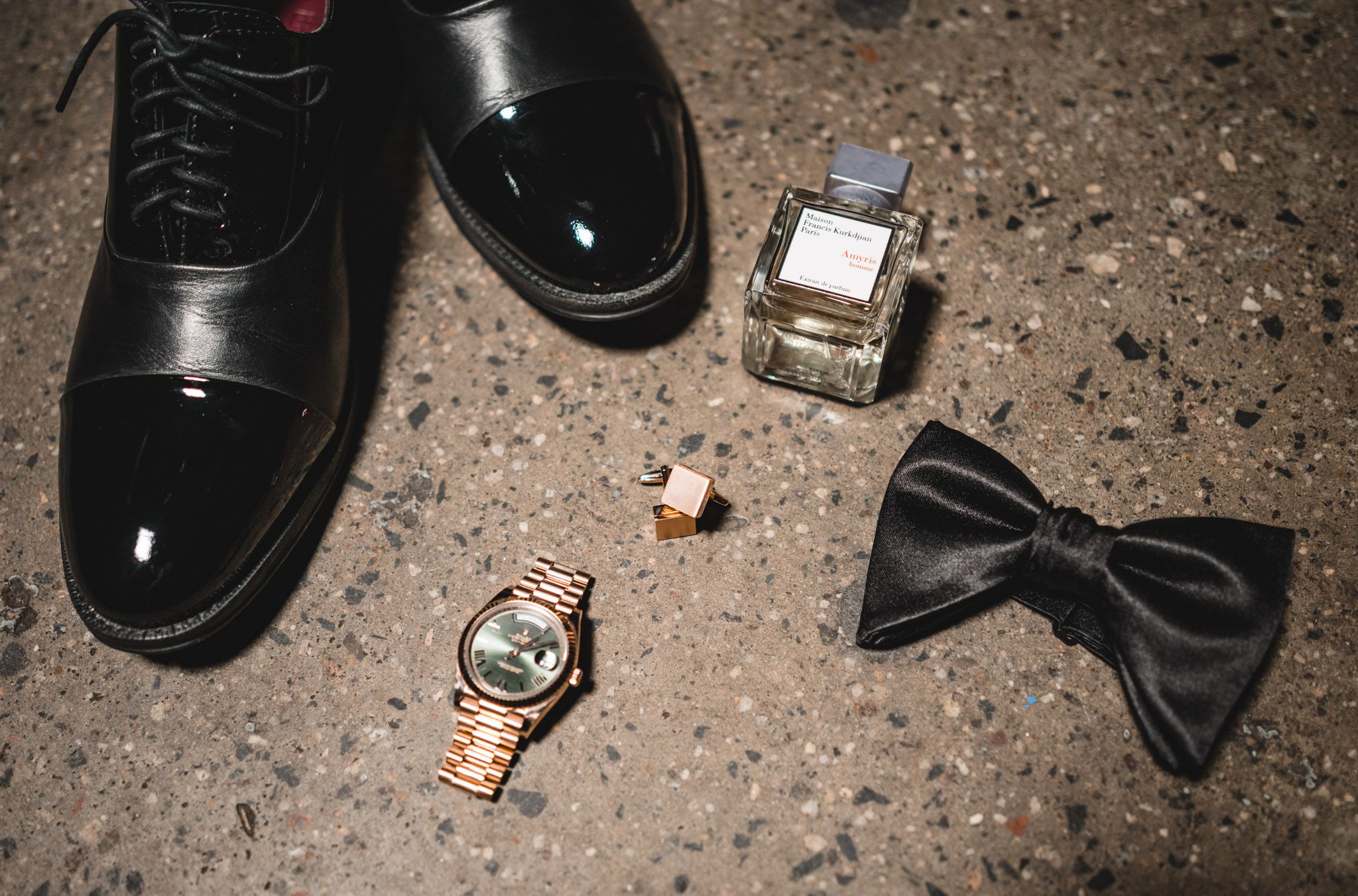 Black patent leather dress shoes, gold Rolex watch, bow tie, cologne, and cufflinks arranged on concrete floor.