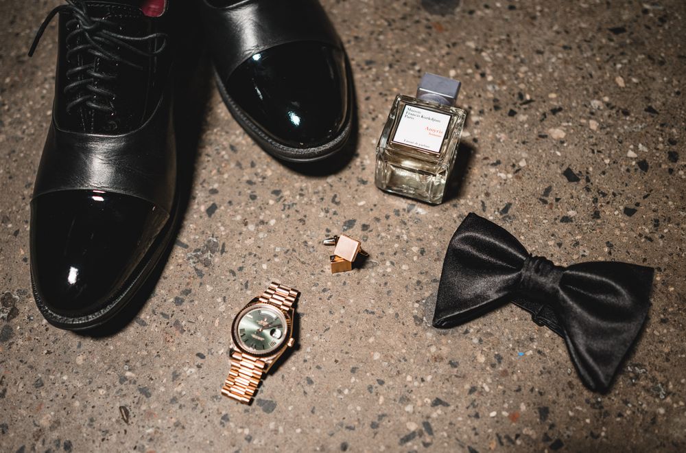 Black patent leather dress shoes, gold Rolex watch, bow tie, cologne, and cufflinks arranged on concrete floor.