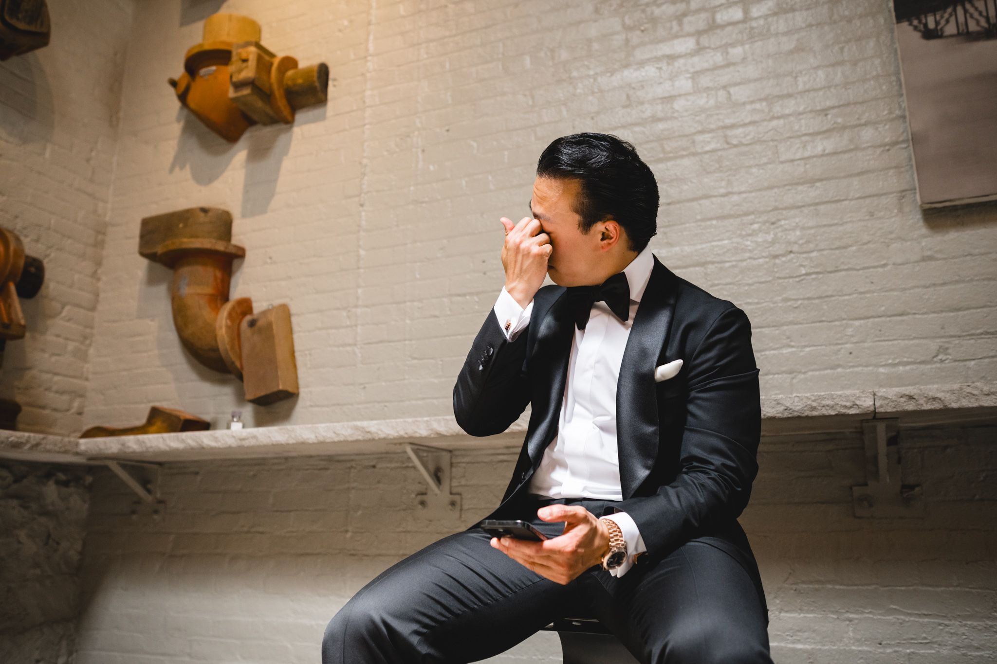 Groom in black tuxedo wiping tears of joy while sitting alone before wedding ceremony.
