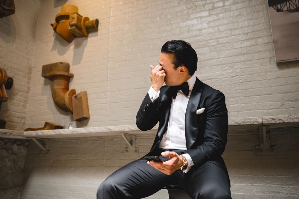 Groom in black tuxedo wiping tears of joy while sitting alone before wedding ceremony.