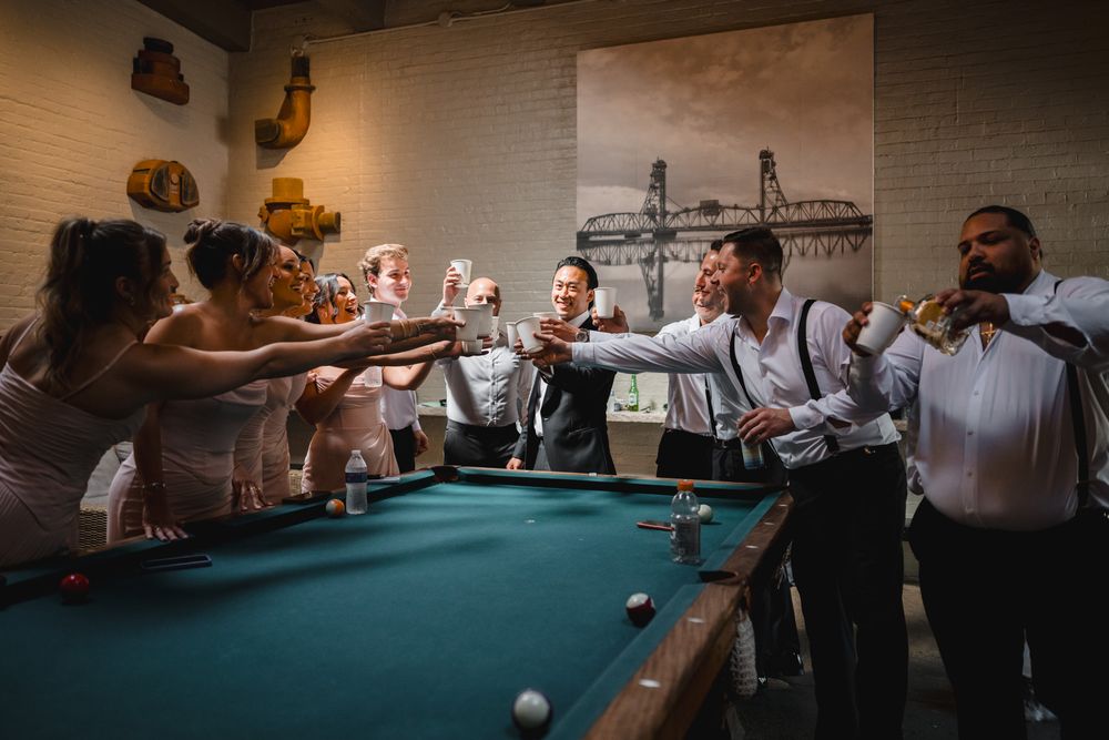 Wedding party celebrating around a pool table, raising glasses in a toast inside a rustic brick-walled venue.