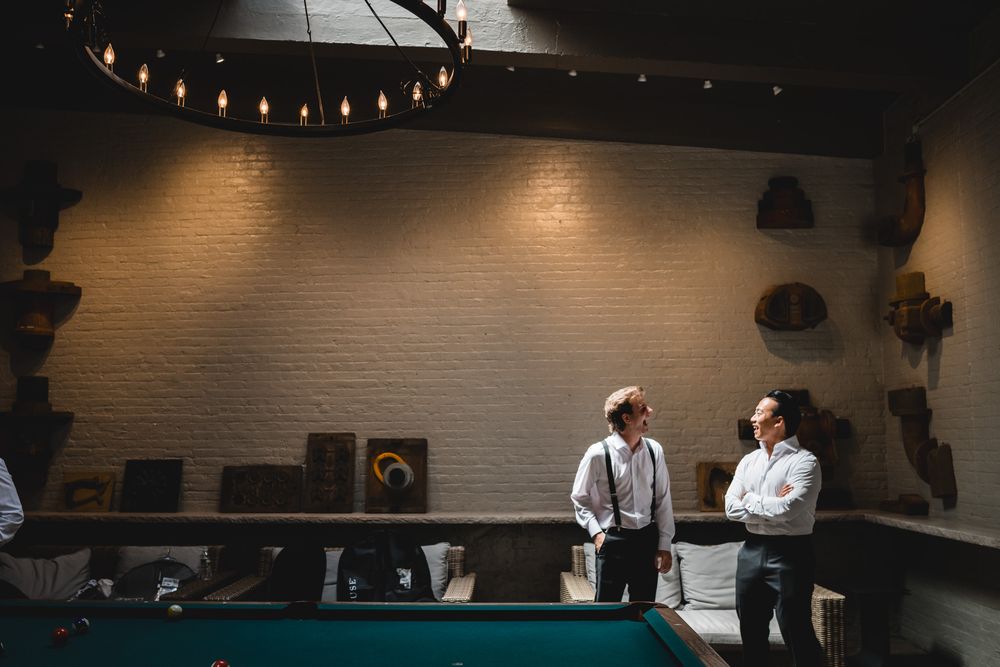 Two groomsmen in suspenders chat beside a pool table in a rustic brick-walled room with Edison bulb lighting.