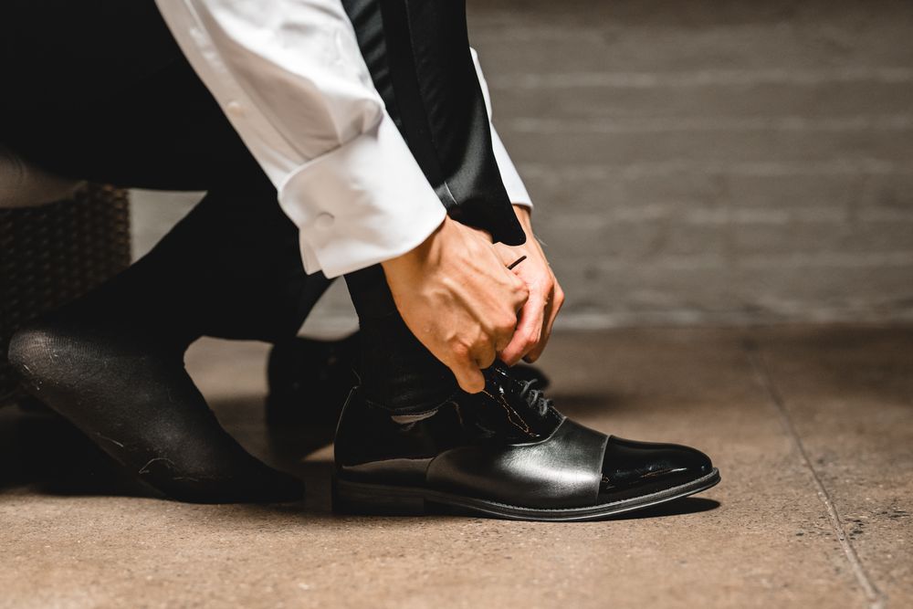 Man in formal suit tying black oxford dress shoes, close-up of hands fastening laces on elegant leather footwear.
