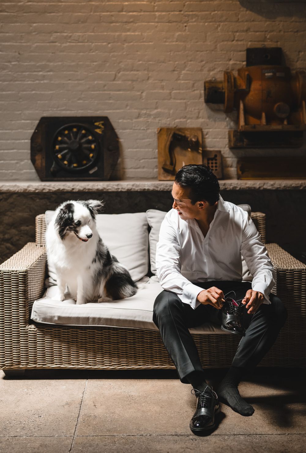 A man in a white shirt sits on a wicker sofa with a border collie dog in a rustic interior setting.