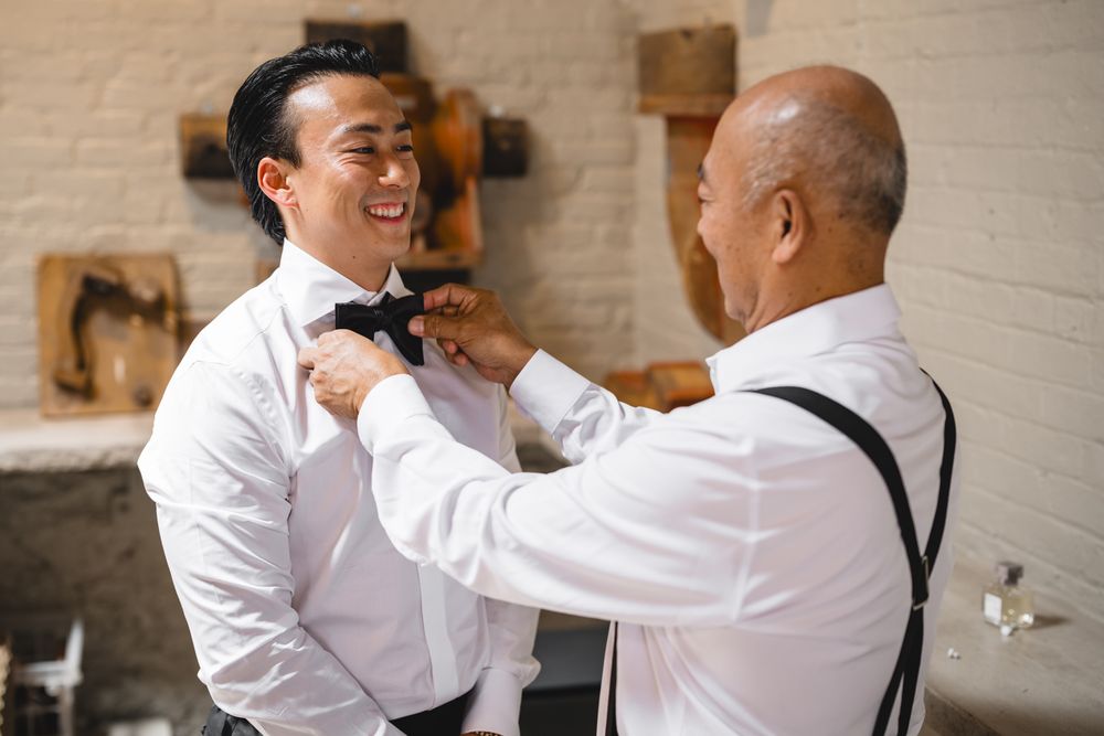 Two men in white dress shirts, one adjusting the other's black bow tie, smiling during wedding preparations.
